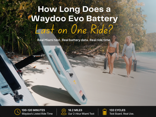 two women walking on a tropical beach carrying eFoil batteries, with Waydoo boards in the foreground and ocean in the background.