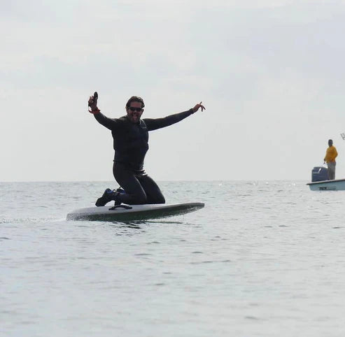 Adult rider in a wetsuit kneeling on an eFoil, smiling with arms outstretched while gliding over calm water in Miami.